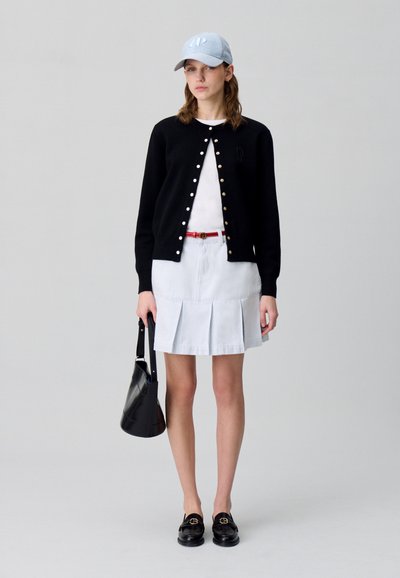 Cardigan nero con bottoni dorati sopra una camicia bianca, gonna plissettata azzurra chiara, cintura rossa, mocassini neri e un cappellino da baseball azzurro chiaro.