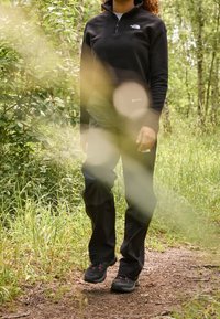 Schwarzer Fleece-Pullover mit einem Reißverschlusskragen, kombiniert mit schwarzen wasserdichten Hosen und robusten schwarzen Wanderschuhen mit roten Akzenten, in einer natürlichen Umgebung.