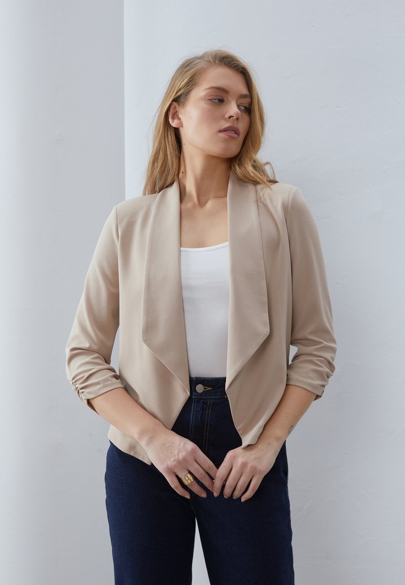 Junge Frau trägt einen beigen Blazer mit offenem Vorderteil, weißes Top und dunkle blaue Jeans, steht an einer hellgrauen Wand.