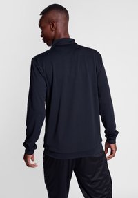 Schwarzes Langarmhemd mit hohem Kragen, strukturiertem Stoff und eng anliegenden Manschetten. Trägt eine Person, die abgewandt steht, kombiniert mit schwarzen Hosen.