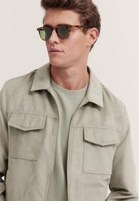Light grey suede jacket with front pockets and a zip closure, worn over a light green t-shirt; paired with tortoiseshell sunglasses.