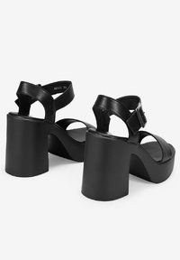 Sandalias de tacón de cuero negro con un tacón grueso, diseño de punta abierta, correa de tobillo ajustable y textura suave. Detalle de hebilla en la correa.