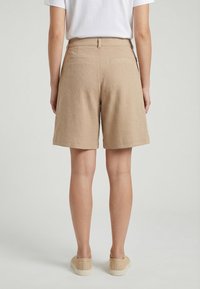 Mujer con pantalones cortos beige texturizados hasta la rodilla, camiseta blanca y zapatos beige sin cierre, de pie de espaldas sobre un fondo liso.