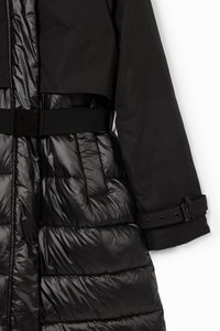 Veste noire avec un haut court et structuré et une partie inférieure matelassée et rembourrée. Comprend une ceinture et des poignets boutonnés. Texture lisse sur l'ensemble.