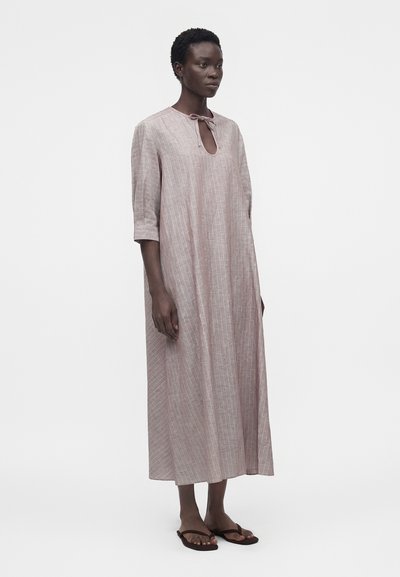 Γυναίκα που φορά χαλαρό, ανοιχτό ροζ φόρεμα μήκους μέχρι τη γάμπα με τριών τετάρτων μανίκια και λαιμόκοψη με άνοιγμα, δεμένη με φιόγκο, όρθια ξυπόλητη σε λευκό φόντο.