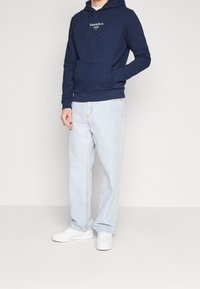 Námořnická mikina s logem "Tommy Jeans 1985", v kombinaci se světle modrými volně střiženými džíny a bílými teniskami. Jednoduchý design a měkká textura.