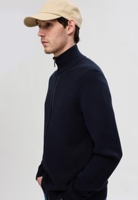 Maglione a zip di colore blu navy con colletto alto, caratterizzato da un motivo testurizzato e una cerniera argentata. Indossato con un cappellino da baseball color beige.