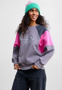 Barevná mikina se šedým tělem a růžovými a černými rukávy, s bílým nápisem "I SOLEMNLY SWEAR I AM UP TO NO GOOD". Doplněná zelenou a růžovou čepicí.