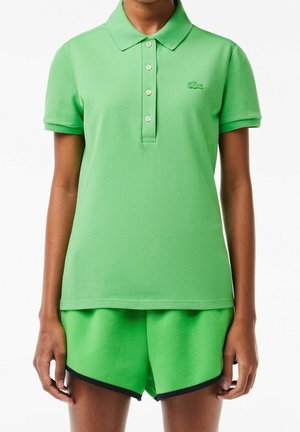Persona que lleva una camiseta polo de manga corta de color verde brillante con botones y pantalones cortos verdes a juego con ribete negro sobre fondo blanco.