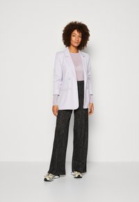 Lys lavendel dobbeltspent blazer over en gjennomsiktig topp, kombinert med blanke svarte vide bukser. Modellen har på seg lyse sneakers.