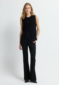 Schwarzes ärmelloses Top mit gerippter Textur und Knopfverschluss, kombiniert mit schwarzen ausgestellten Hosen und spitzen schwarzen Schuhen. Einfaches Design.