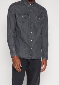 Tummansininen farkkupaita pitkillä hihoilla, joissa on kaksi rintataskua, terävä kaulus ja valkoiset nepparit. Tyylissä on hienovarainen tekstuuripinta.