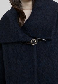 Granatowy wełniany płaszcz z dużym kołnierzem, ozdobiony błyszczącą czarną klamrą w talii, z miękkim, teksturowanym wykończeniem.