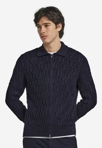 Marinblå stickad cardigan med en texturerad diamantmönster, dragkedja fram och ribbad krage och ärmslut. Tillverkad av ett mjukt, stretchigt material.