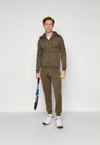 Mladý muž v hnedom športovom oblečení, ktorý drží tenisovú raketu a tenisovú loptu, stojí na jednoduchom bielom pozadí.