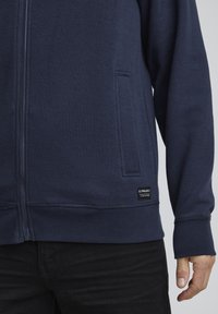 Mørkeblå zip-up hettegenser laget av myk stoff, med en sidelomme, ribbestrikkede mansjetter og et merke på kanten.