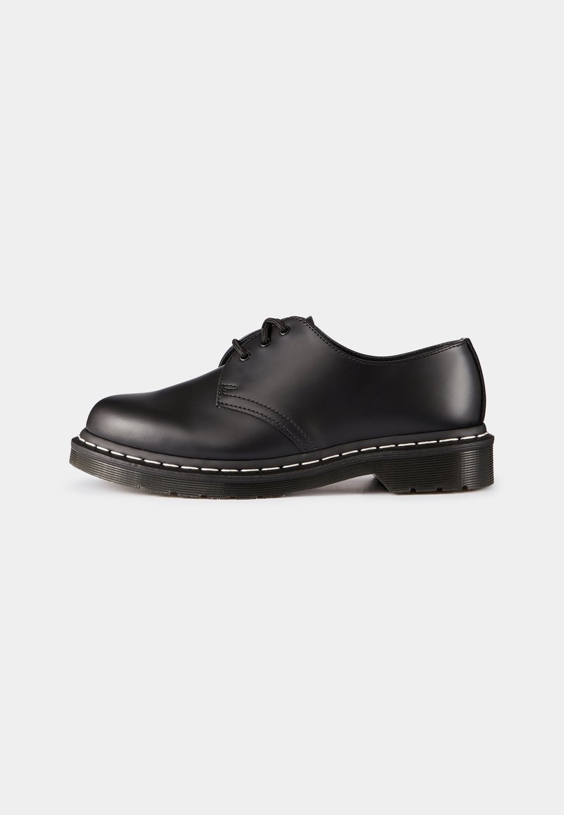 Chaussure oxford basse en cuir noir avec trois œillets, détails de couture blanche et semelle en caoutchouc épaisse sur un fond gris clair.