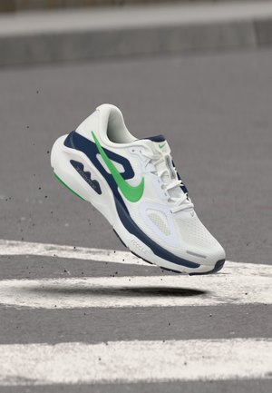 Bielo-modrá bežecká obuv so zeleným logom Nike, zachytená vo vzduchu nad sivou cestou s bielymi značkami.