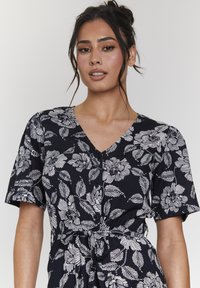 Vestido de envoltura floral en color marino, con estampado de flores y hojas en blanco, mangas cortas, escote en V y detalle de cintura anudada. Textura de tejido suave.