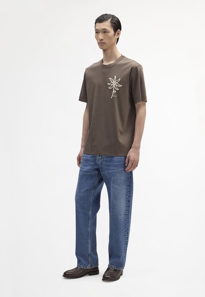 Bruine katoenen t-shirt met korte mouwen, voorzien van een beige bloemborduurwerk op de borst, gecombineerd met losse blauwe denim jeans en donkere schoenen.