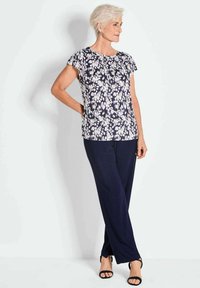 Bloemige top met korte mouwen en marineblauwe achtergrond, met witte en lila bloemen. Gekleed met donkere blauwe broeken en zwarte sandalen.