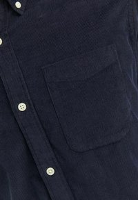 Camisa de pana en color azul marino con estrías verticales, un bolsillo en el pecho y botones blancos a lo largo de la botonadura. La tela tiene una textura suave.