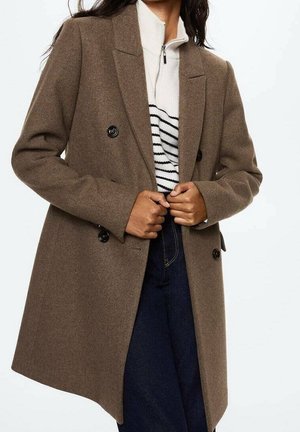 Kvinde iført en brun dobbeltbrystet frakke, stribet hvid lynet sweater og mørkeblå jeans, der holder i frakkens slag.