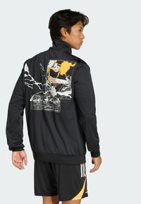 Young man wearing a black jacket with a colorful graphic design on the back, paired with black and yellow sports shorts.