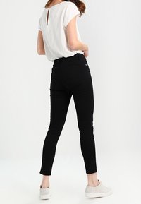 Jeans ajustados negros combinados con una blusa ligera de manga corta con un detalle en forma de ojal en la parte posterior y zapatos blancos tipo slip-on. Fondo liso.