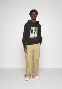 Felpa nera con stampa grafica, caratterizzata da dettagli bianchi, abbinata a pantaloni cargo beige e sneakers bianche. Il modello si trova davanti a uno sfondo semplice.
