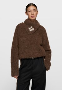 Suéter de punto marrón y esponjoso con un cuello alto, con una franja diagonal blanca. Combinado con pantalones negros de corte holgado.