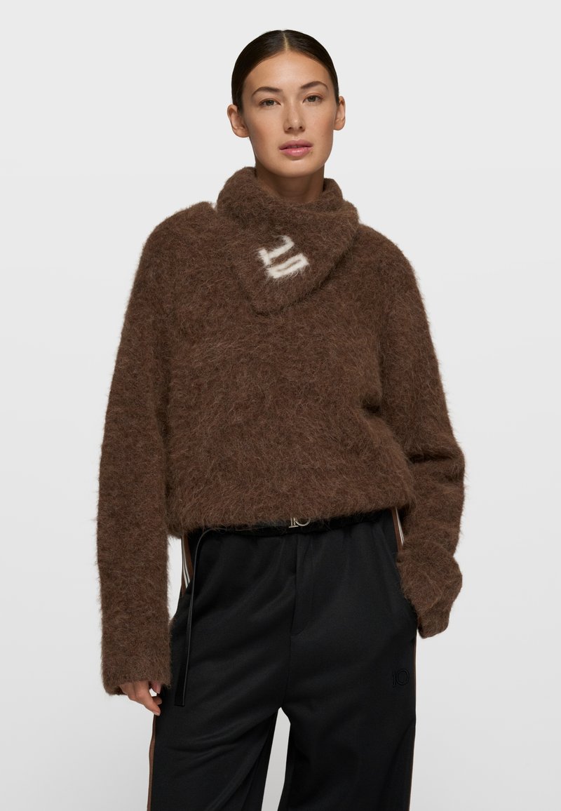 Suéter de punto marrón y esponjoso con un cuello alto, con una franja diagonal blanca. Combinado con pantalones negros de corte holgado.