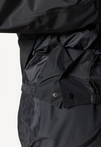 Schwarze wasserdichte Jacke mit einer glatten, matten Textur. Verfügt über eine taillierte Taille, Druckknopfverschlüsse und ein dezentes gestreiftes Muster an der Seite.