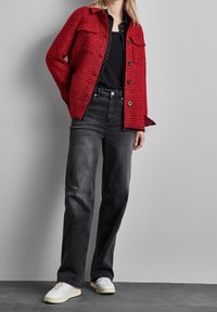 Red textured jacket with two chest pockets, paired with high-waisted black jeans and white sneakers. Smooth surface and relaxed fit.