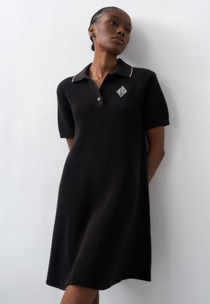 Kvinde iført en sort, kortærmet, ribstrikket polo-kjole med en hvid logo på venstre bryst, stående mod en ensfarvet lys væg.