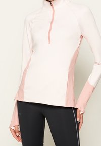 Světle růžová sportovní mikina na zip s růžovými bočními panely, dlouhými rukávy a otvory na palce, vyrobená z elastického materiálu. Spárována s černými legínami.