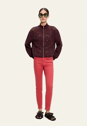 Kobieta stojąca, nosząca duże, okrągłe okulary przeciwsłoneczne, ciemnofioletową szydełkową kurtkę na zamek, koralowe obcisłe spodnie oraz czarne sandały z frędzlami.