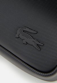 Svart teksturert etui med et hevet svart krokodillelogo, har en glatt kant og en slitesterk finish. Egnet for kompakt lagring.