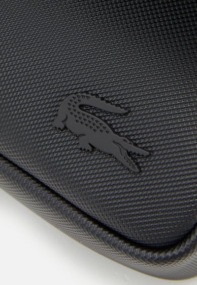 Étui noir texturé avec un logo de crocodile noir en relief, présente un bord lisse et une finition durable. Convient pour un rangement compact.