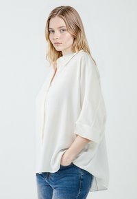 Loose-fitting white blouse with a button-up front, rolled sleeves, and soft fabric. Complemented by light denim jeans for a casual look.