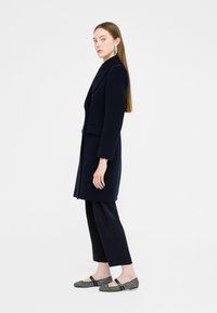 Marineblauwe lange jas met geknipte kraag, zwarte broek en gemusterde ballerina's met een band en gespsluiting. Model staat in profiel tegen een witte achtergrond.