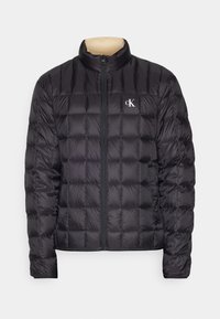 Veste matelassée noire avec fermeture éclair, doublure intérieure beige au col et logo "cK" blanc sur la poitrine gauche, manches longues et col montant.