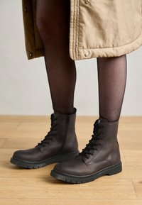 Botas de cuero marrón hasta el tobillo con cordones, una superficie texturizada y una suela de goma negra y resistente. Combinadas con medias negras transparentes y un abrigo beige.