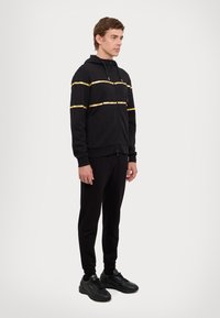 Black zip-up hoodie with gold horizontal stripes, paired with black joggers and sneakers. Features a hood and side pockets.