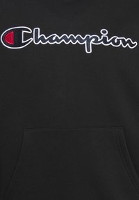 Tissu noir avec logo Champion blanc et rouge brodé au-dessus d'une poche kangourou avant sur un sweat-shirt ou un sweat à capuche.