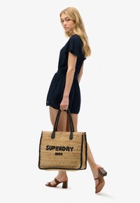 Woven straw tote bag med sorte læderhåndtag, prydet med "SUPERDRY IBIZA" i fed tekst. Tasken har en rektangulær form og dekorative detaljer.