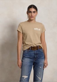 Beżowa bawełniana koszulka z napisem "POLO" w kolorze białym. Noszona z podartymi niebieskimi dżinsami i brązowym paskiem, z widocznymi przeszyciami i detalami metalowymi.