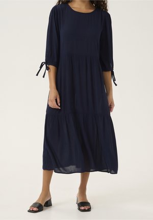 Kvinde iført en navyblå midi-kjole med 3/4-ærmer, der er bundet ved manchetten, kombineret med sorte sandaler med blokhæl.