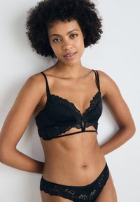 Črn komplet spodnjega perila, ki vključuje bralette iz spuščene čipke z tankimi naramnicami in ujemajoče se čipkaste hlačke, ki poudarjajo eleganten material in odprt hrbtni del.