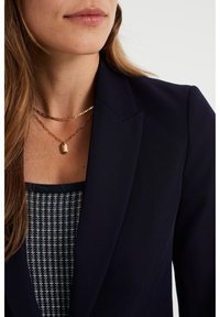 Navy blazer with notched lapel over a patterned top; layered gold necklaces, one with a rectangular pendant. Smooth texture and tailored fit.